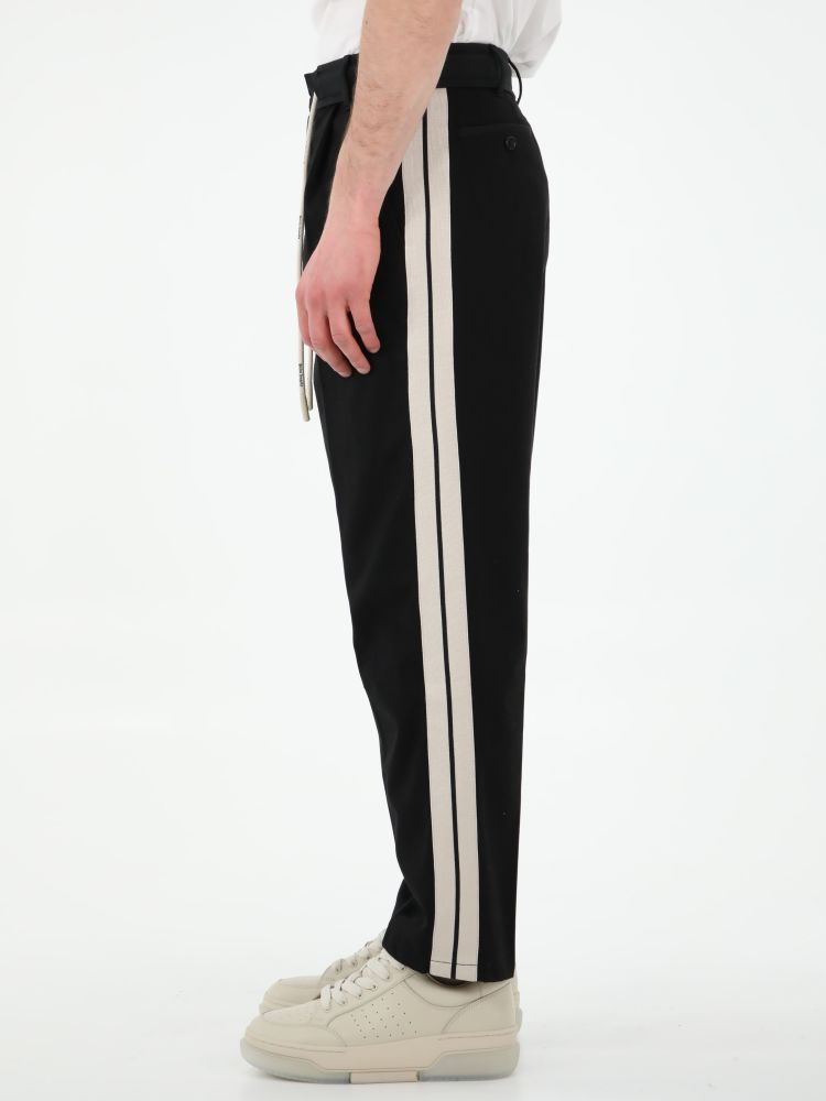 Black track pants with tone-on-tone belt and white drawstring with Palm Angels logo. It features white side stripes, two side welt pockets, two rear buttoned welt pockets and belt loops. The model is 184cm tall and wears size 48.