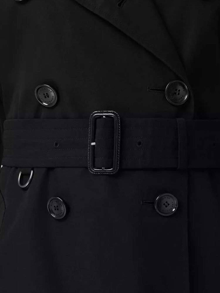 Kensington Heritage trench coat in black cotton gabardine with double-breasted closure and adjustable belt on waist. It features epaulettes, hook-and-eye collar closure, two side buttoned welt pockets and belted cuffs. The model is 179cm tall and wears size UK 6.
