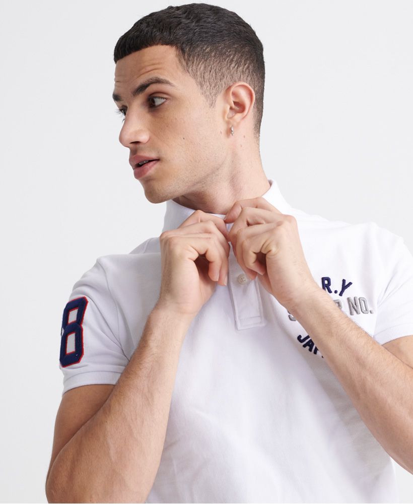 Superdry men's Classic superstate polo shirt. A staple in any wardrobe, this short sleeved polo features a button fastening at the collar, side splits at the hem, and a longer hem at the back. Finished with an embroidered number on one sleeve, and an embroidered Superdry logo on the chest.Made with Organic Cotton - Grown using only organic inputs and no artificial chemicals, which leads to improved soil condition, stronger biodiversity and better health among the cotton growers and uses between 60-90% less water to grow. By 2030, all Superdry Cotton will be Organic.Slim fit