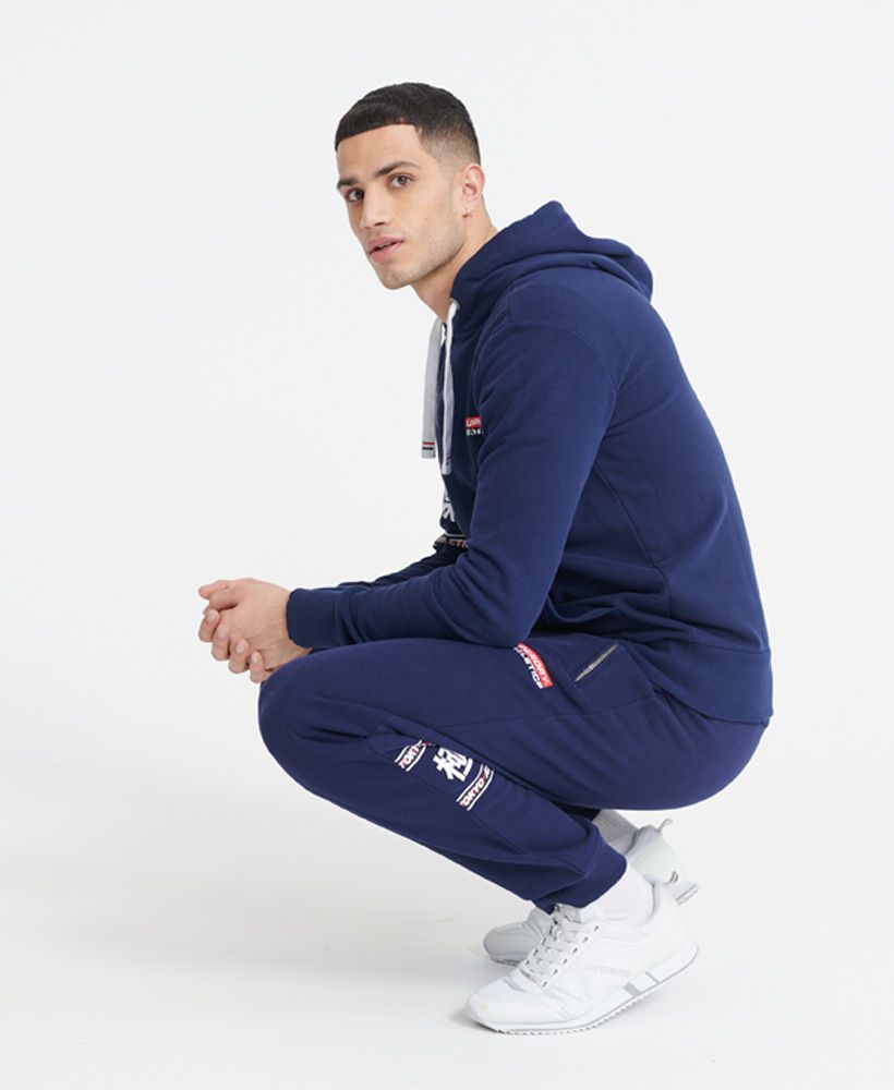 Superdry men's Trophy lightweight joggers. Featuring an elasticated drawstring waist, two front zipped pockets, and cuffed ribbed cuffs. Finished with a small Superdry logo print on the front, and a large Superdry dry print with Japanese characters on the back of one leg.