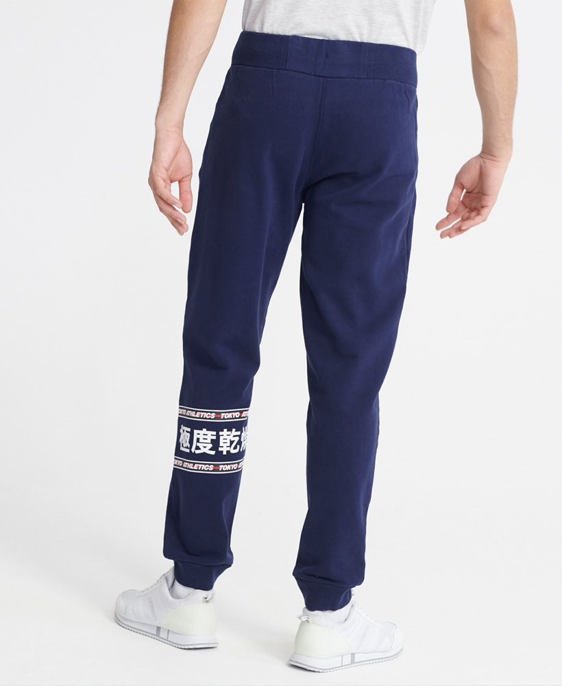 Superdry men's Trophy lightweight joggers. Featuring an elasticated drawstring waist, two front zipped pockets, and cuffed ribbed cuffs. Finished with a small Superdry logo print on the front, and a large Superdry dry print with Japanese characters on the back of one leg.