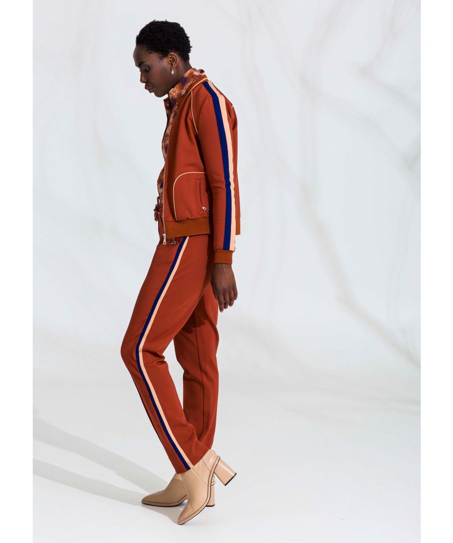 Bruine broek met smalle pijpen met elastische tailleband met koord om de broek aan te trekken De broek heeft witte en rode strepen aan de zijkanten en zakken.